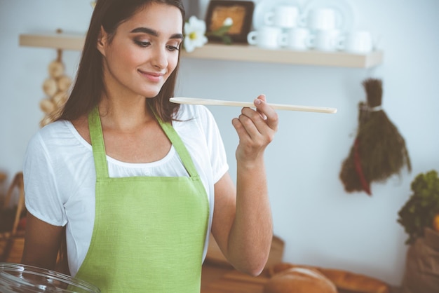 Una atractiva joven de cabello oscuro que prueba comida caliente lista con una cuchara de madera mientras está de pie y sonríe cerca de la estufa de la cocina Conceptos de cocina y hogar