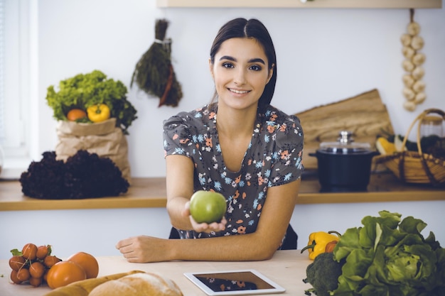 Foto una atractiva joven de cabello oscuro sosteniendo una manzana verde y eligiendo la receta de una deliciosa comida mientras sonríe en la cocina tablet pc es el mejor libro de cocina