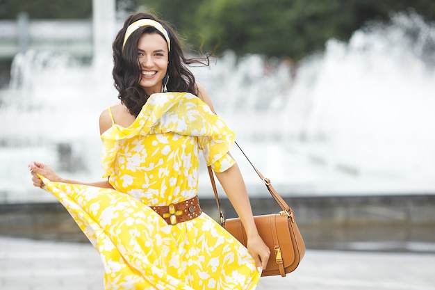 Atractiva joven caminando por la ciudad. Señora de vestido largo amarillo cerca de la fuente. Hermosa mujer sobre fondo de verano.