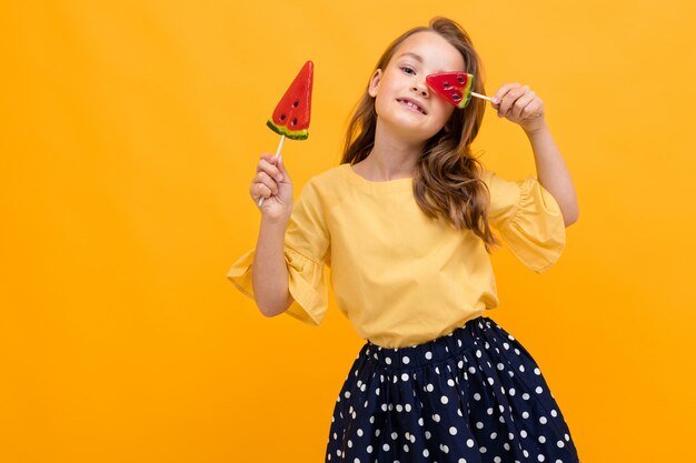 Atractiva joven caucásica en una falda y una camiseta con dos paletas de sandía sobre un fondo amarillo