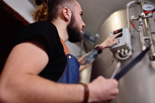 Una atractiva joven cervecera con barba examina el equipo para elaborar cerveza y registra los resultados en papel.