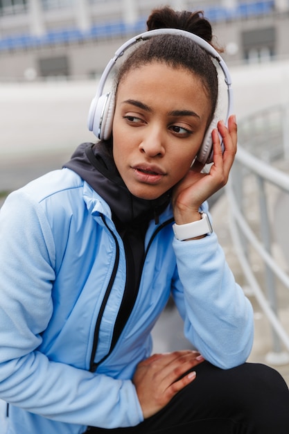 Atractiva joven deportista africana sana escuchando música con auriculares inalámbricos escurriendo en el estadio