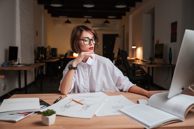 Atractiva joven diseñadora de moda en gafas sentado y trabajando en la oficina