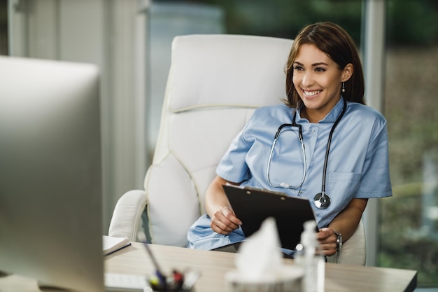 Una atractiva joven doctora sentada en su consultorio y mirando la computadora.