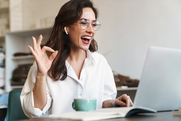 Atractiva joven empresaria morena alegre sentada en la mesa de café con computadora portátil en el interior, usando auriculares inalámbricos, llamada de audio, gesto ok