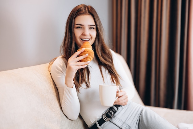 Atractiva joven está comiendo croissant y tomando café en casa