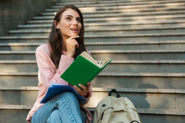 Atractiva joven estudiante mujer sonriente leyendo un libro mientras está sentado en la escalera