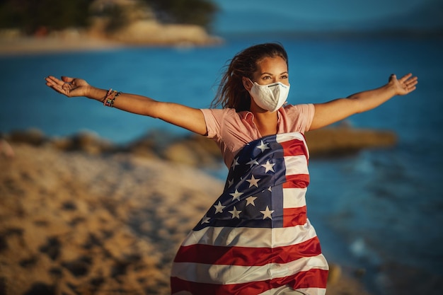 Atractiva joven feliz con bandera nacional estadounidense y máscara protectora N95 en la playa durante la prevención de la propagación de la epidemia y el tratamiento del coronavirus y la pandemia COVID-19.