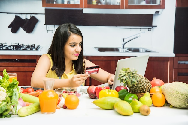 Atractiva joven india con delantal en la cocina de compras en línea con tarjeta de débito o crédito en la computadora portátil con mesa llena de frutas y verduras frescas