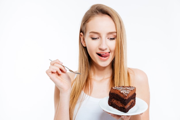 Atractiva joven juguetona sosteniendo y comiendo un trozo de pastel de chocolate sobre fondo blanco.