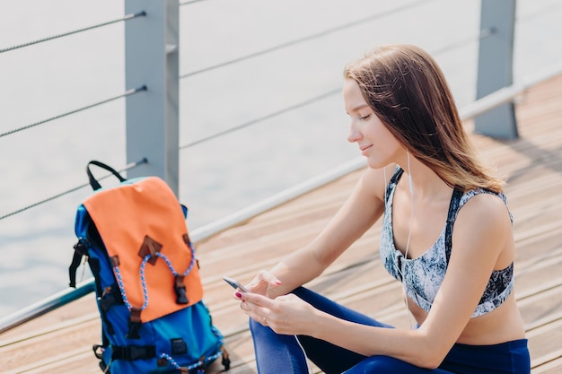 Atractiva joven y linda deportista con top informal que tiene una figura perfecta que usa un teléfono inteligente moderno para escuchar música y enviar mensajes revisa los tipos de buzones de correo electrónico poses de comentarios al aire libre cerca de la mochila