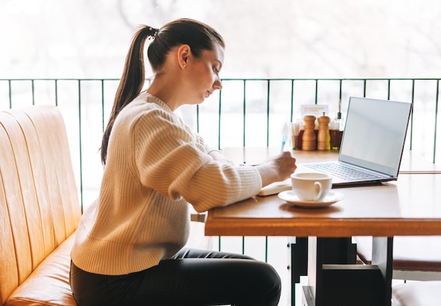 Atractiva joven morena sonriente estudiante independiente con una taza de té usando una computadora portátil en el café