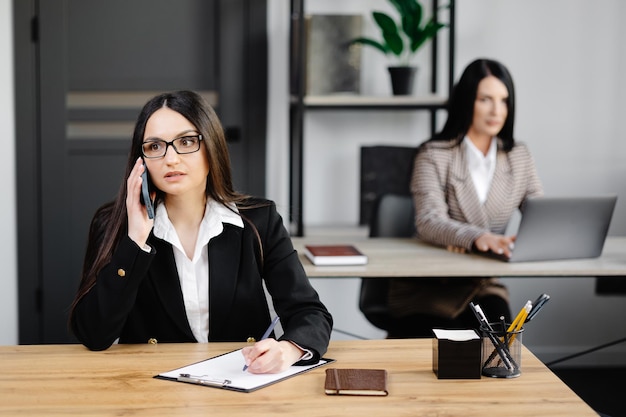 Atractiva joven mujer de negocios con chaqueta hablando por teléfono móvil mientras se sienta en un escritorio y usa un cuaderno cerca de su colega en el trabajo en la oficina