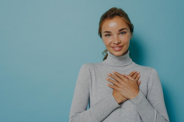 Atractiva joven mujer sonriente sosteniendo las manos juntas cerca del corazón en el pecho mientras posa en el estudio