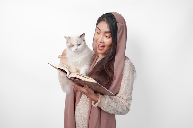 Atractiva joven musulmana asiática con velo y hijab sonriendo mientras abraza a una muñeca blanca y sostiene el Corán por otro lado