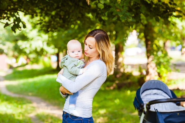 Atractiva joven parada cerca de un cochecito de bebé en el parque de verano con su hijo en brazos. Concepto de familia feliz