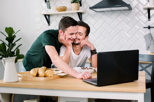 Atractiva joven pareja caucásica gay, pareja homosexual, en la cocina, abrazándose y desayunando en la mesa, usando internet en la computadora portátil. Estudio, cocina ligera de fondo.