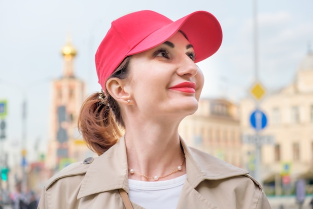 Atractiva joven pelirroja con una gorra de béisbol roja y un impermeable beige se encuentra en el contexto de los edificios de la ciudad y mira hacia arriba. Retrato de primer plano de una mujer sonriente. Caminando por la ciudad.