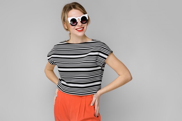 Atractiva joven rubia en blusa a rayas sonriendo en gafas de sol con las manos en la cintura en gris