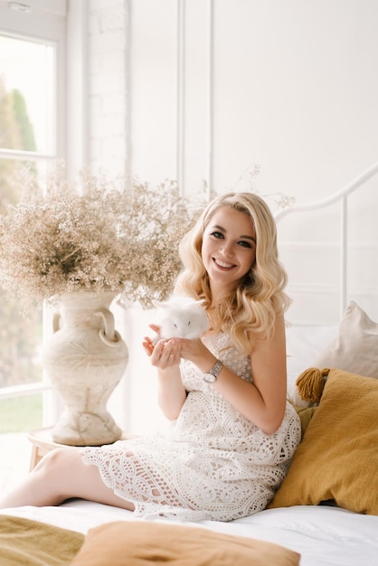 Atractiva joven rubia con cabello rizado y maquillaje con un vestido blanco con un conejo en las manos en el luminoso interior de la casa estudio