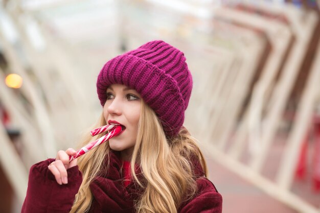 Atractiva joven rubia con gorro de punto rojo posando con bastón de caramelo en la calle