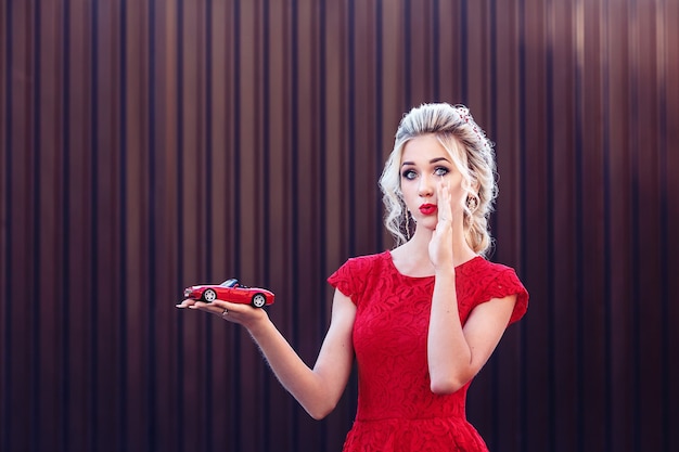 Atractiva joven rubia con un vestido rojo sosteniendo un coche de juguete. Concepto de arrendamiento y compra de un automóvil