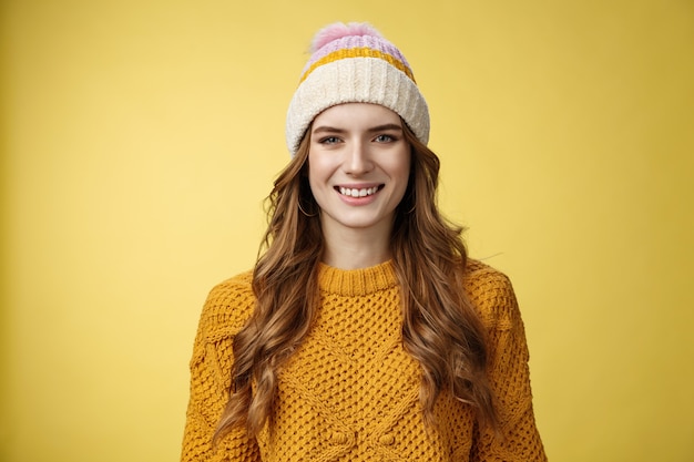 Atractiva joven saliente de aspecto amistoso vestida de viaje cálido montañas esquiando divirtiéndose pasar las vacaciones de invierno en los Alpes en familia, sonriendo ampliamente con suéter de sombrero de pana, fondo amarillo