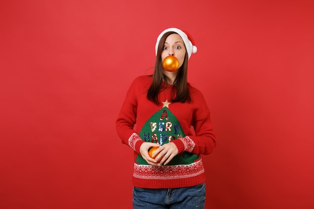 Atractiva joven Santa en suéter, sombrero de Navidad con juguetes de árbol amarillo en la boca y manos aisladas sobre fondo rojo. Feliz año nuevo 2019 celebración concepto de fiesta navideña. Simulacros de espacio de copia.