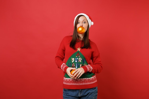 Foto atractiva joven santa en suéter, sombrero de navidad con juguetes de árbol amarillo en la boca y manos aisladas sobre fondo rojo. feliz año nuevo 2019 celebración concepto de fiesta navideña. simulacros de espacio de copia.