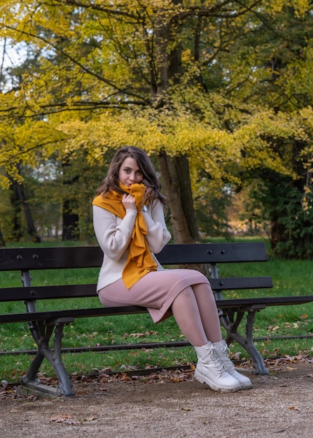 Atractiva joven sonriente sentada en un banco en un parque de otoño envuelta en un chal amarillo