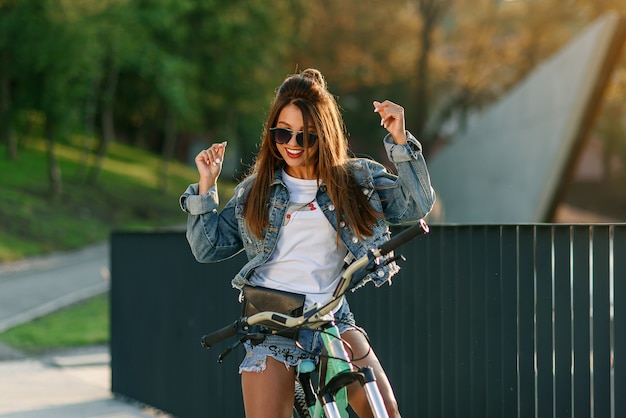 Foto atractiva joven tentadora en ropa de jeans de moda usa gafas con estilo, sentada en la bicicleta y posando ante la cámara en la noche de verano. camara lenta