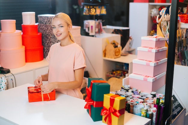 Atractiva joven vendedora sosteniendo y usando cinta dorada para atar el lazo para la caja de regalo envuelta en rojo para ...