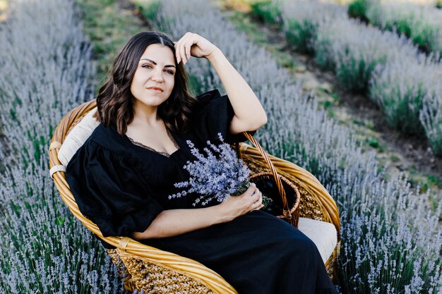 Atractiva joven vestida de negro sentada en una silla rodeada de un campo de lavanda y sosteniendo una cesta con flores