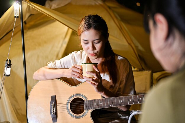 Atractiva joven viajera asiática bebiendo un chocolate caliente mientras disfruta de una fiesta nocturna de campamento