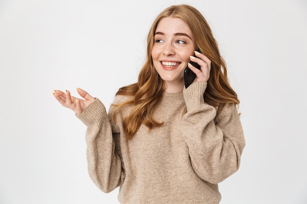 Atractiva joven vistiendo suéter que se encuentran aisladas sobre una pared blanca, hablando por teléfono móvil