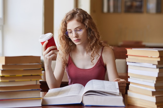 Atractiva jovencita con gafas y uotfit casual, leyendo el libro que pone en la mesa y sosteniendo la taza de café en la mano mientras está sentado cerca del escritorio de madera con pila de libros, cerca de la ventana.