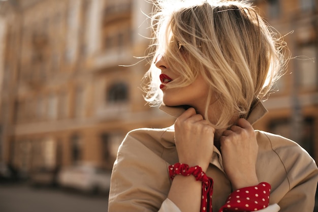 Atractiva jovencita rubia con poses de labios rojos en una gabardina de moda beige fuera de la mujer con gafas de sol mira hacia el exterior