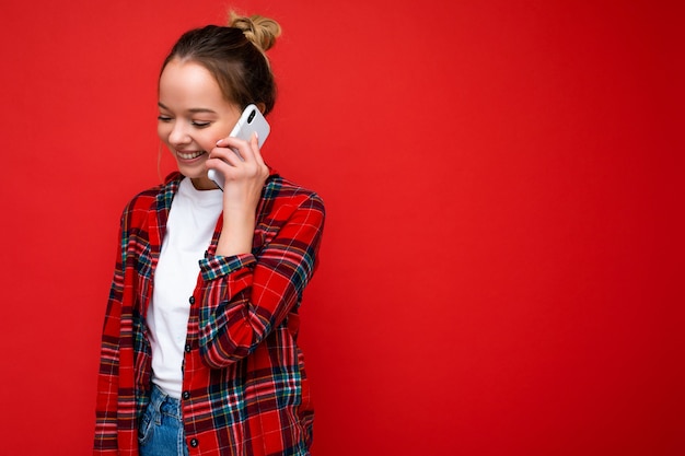 Atractiva linda mujer rubia joven positiva que se encuentran aisladas sobre la pared roja vistiendo casual camisa roja y camiseta blanca hablando por teléfono móvil mirando hacia abajo