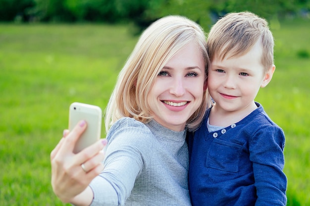 Atractiva madre e hijo amado toman fotos selfie en teléfono en el parque sobre un fondo de hierba verde y árboles.