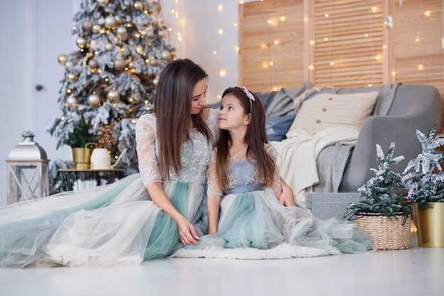 Atractiva madre y su hija linda en un suave vestido de noche sentado en el suelo cerca del árbol de Navidad en una acogedora habitación.