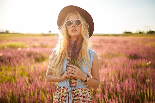 Atractiva modelo femenina con cabello rubio, sombrero y gafas de sol posando en un campo floral detrás del fondo del atardecer