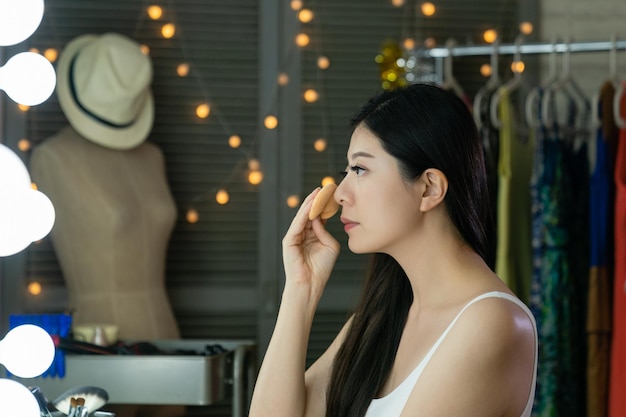 Atractiva modelo llamativa mujer japonesa maquillada frente al espejo con bombillas en el tocador en el backstage. vista lateral de una dama asiática que se mira a sí misma mientras se pone el polvo en la mejilla en el vestidor