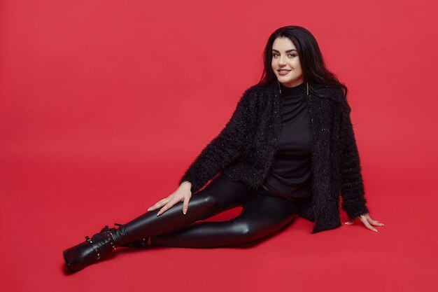 Atractiva morena con cabello largo sentado en el estudio sobre un fondo rojo.