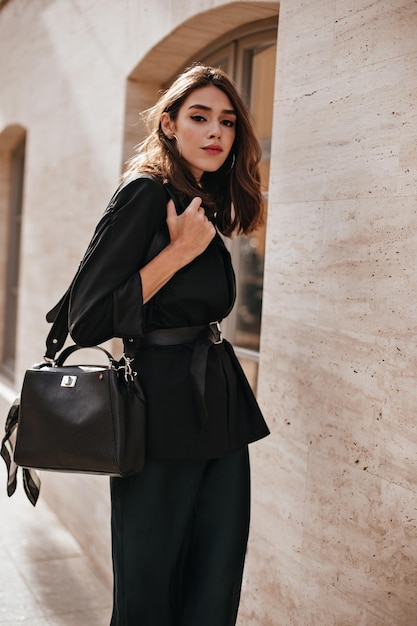 Atractiva morena joven con chaqueta de vestir oscura de maquillaje brillante y accesorios posando al aire libre contra un fondo de pared beige claro y mirando directamente