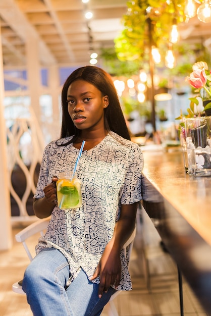 Atractiva mujer africana sentada en la cafetería y bebiendo limonada fresca.