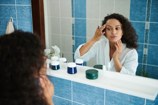 Una atractiva mujer afroamericana de mediana edad examina su rostro y párpados en el reflejo del espejo del baño. Concepto de cuidado de la piel