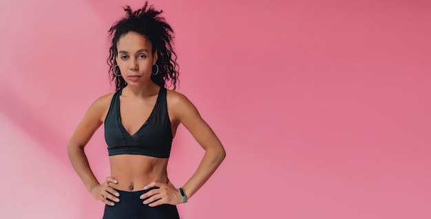 Atractiva mujer afroamericana negra en polainas negras y ropa de fitness superior sobre fondo rosa aislado cabello rizado confiado estilo deportivo cuerpo atlético