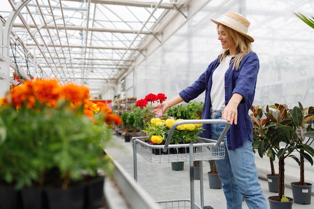 Atractiva mujer alegre de mediana edad con flores en macetas en carrito de compras en invernadero