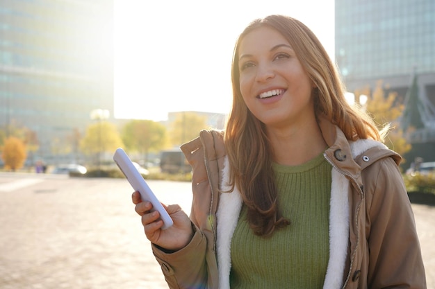 Atractiva mujer alegre sosteniendo un teléfono móvil en la ciudad sonriendo mirando hacia un lado y mirando hacia otro lado pensando