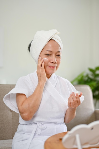 Una atractiva mujer asiática en bata de baño aplicando crema facial haciendo su rutina de cuidado de la piel